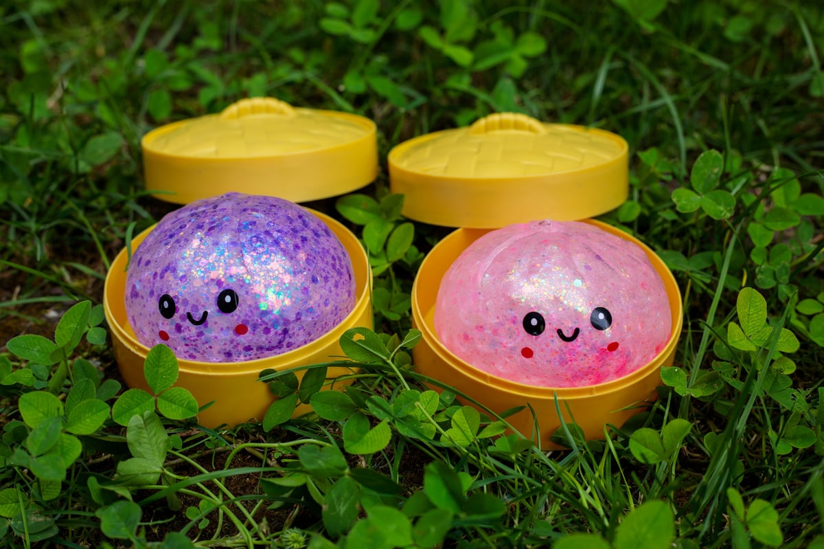 Two kawaii glitter squishy dumpling toys in purple and pink sitting in open yellow steamer containers